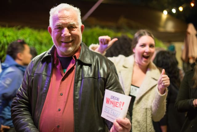 Man in a leather jacket smiling and holding a sign that reads "Raffle Winner!" with a woman in a white coat giving a thumbs-up in a festive outdoor setting at the Love Bomb 2017 event supporting My Voice Music.
